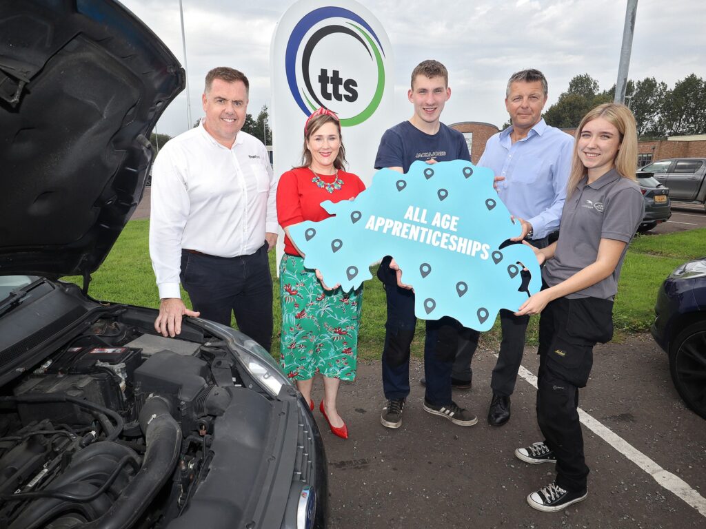 The Department for Economy announcing the All Age Apprenticships with  two apprentices holding an Northern IreIand map cut out that says 'All Age apprentices on it. Surrounded by a manager and a representative from the Department.